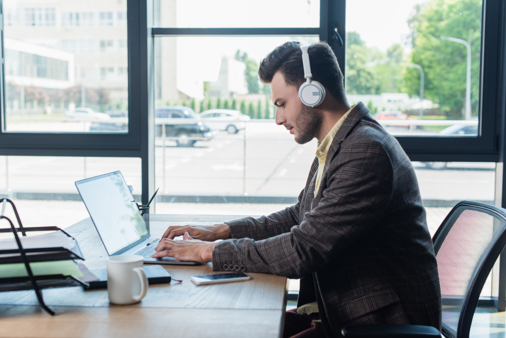 Вид збоку молодого бізнесмена в навушниках з використанням ноутбука біля паперу і чашки в офісі
  - Фото, зображення