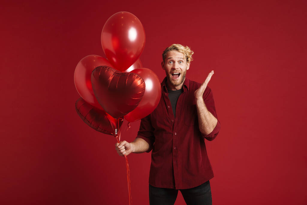 Bearded blonde man expressing surprise while posing with balloons isolated over red background - 写真・画像