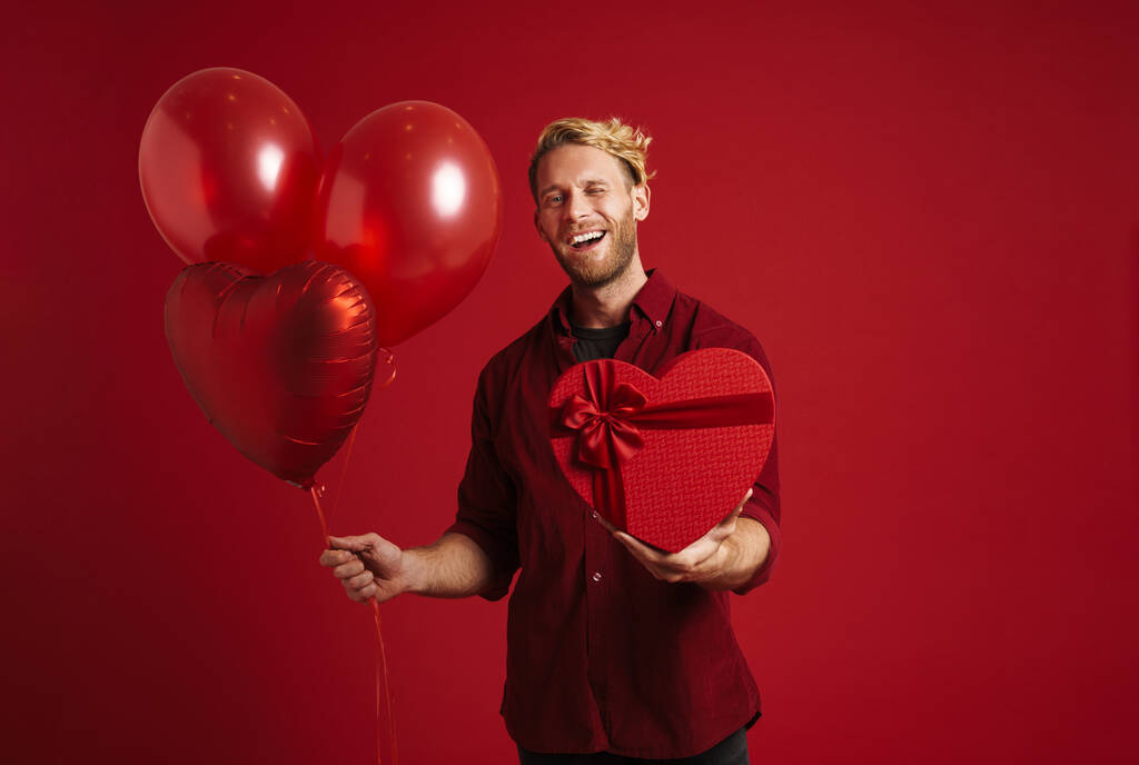 Beyaz adam kırmızı duvardan izole edilmiş balonlar ve kalp hediye kutusuyla poz verirken göz kırpıyor. - Fotoğraf, Görsel