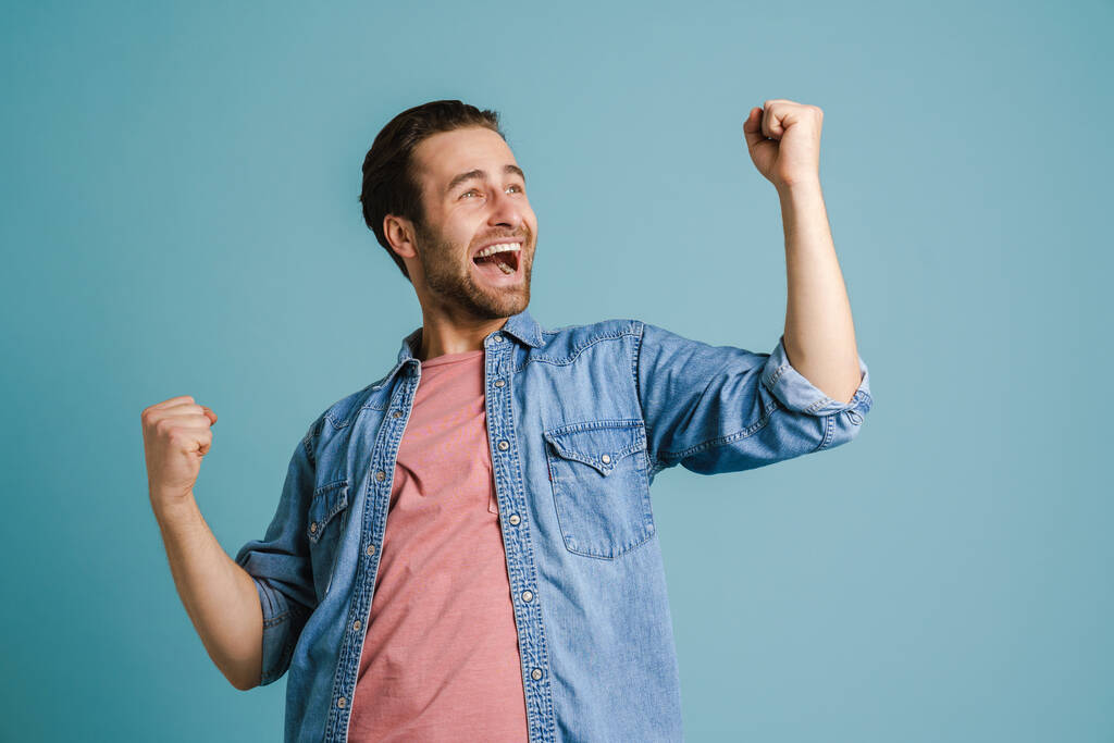 Fiatal izgatott férfi sikoltozik, miközben a győztes gesztus elszigetelt kék háttér - Fotó, kép