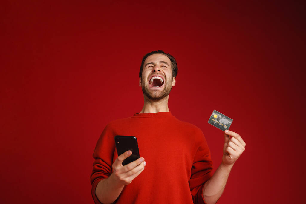 Surprised man screaming while showing credit card and cellphone isolated over red background - Foto, Imagen