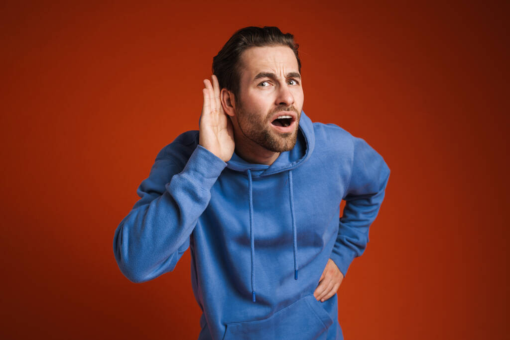 Young man listening and gesturing while expressing surprise isolated over red background - Foto, imagen