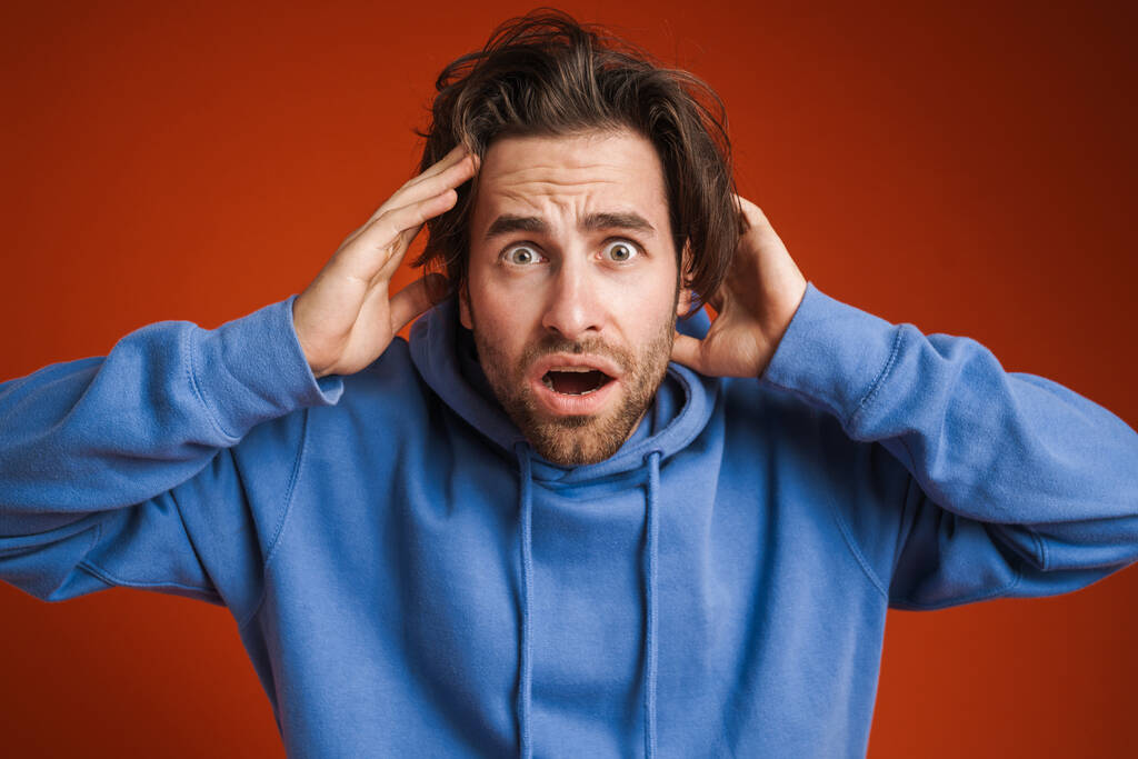 Young man holding his head while expressing surprise isolated over red background - Foto, afbeelding
