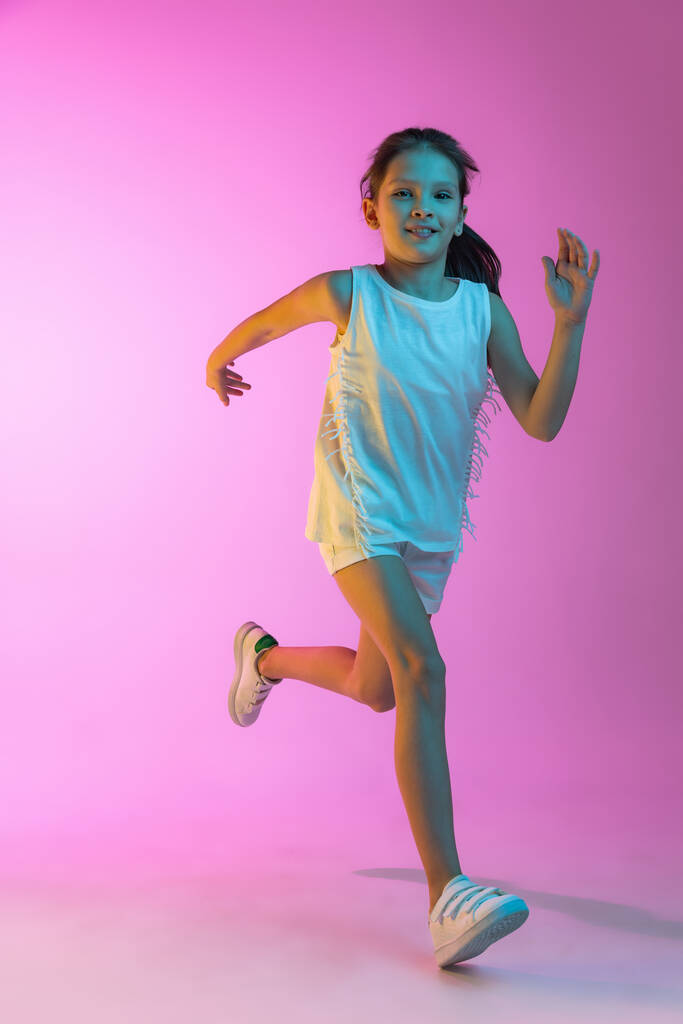 Utíkám. Šťastné malé krásné dítě, školní dívka v ležérní oblečení izolované na růžovém pozadí v neonu. Koncept dětské emoce, móda, krása, zpět do školy a reklama - Fotografie, Obrázek