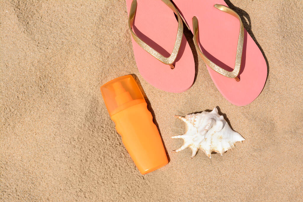 Bottle with sun protection spray, flip flops and seashell on sandy beach, flat lay. Space for text - Photo, image