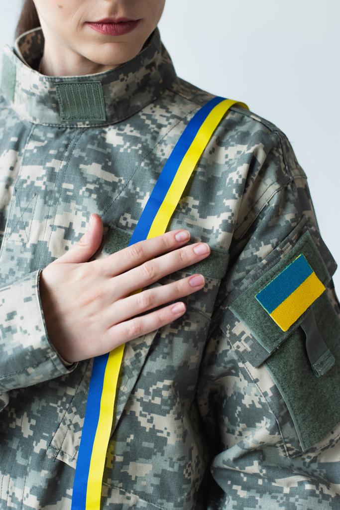 Cropped view of soldier with ukrainian flag on chevron touching blue and yellow ribbon isolated on grey  - Photo, image