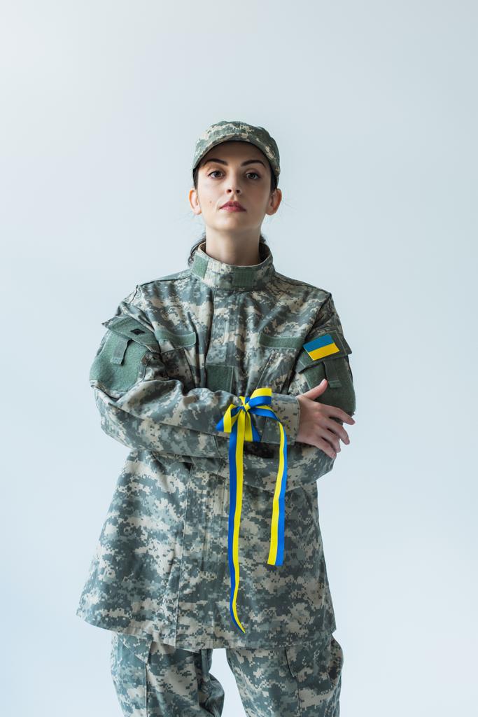 Soldier in uniform with ukrainian flag and blue and yellow ribbon crossing arms isolated on grey  - 写真・画像