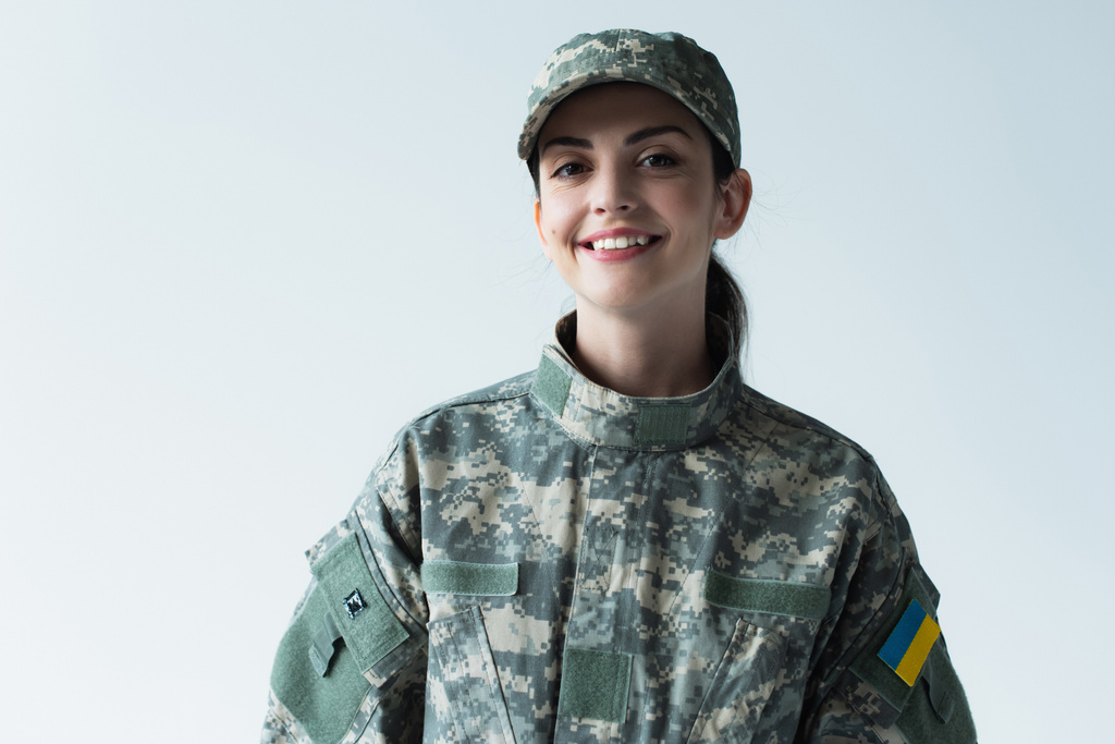 Cheerful soldier in uniform with ukrainian flag looking at camera isolated on grey  - Photo, Image