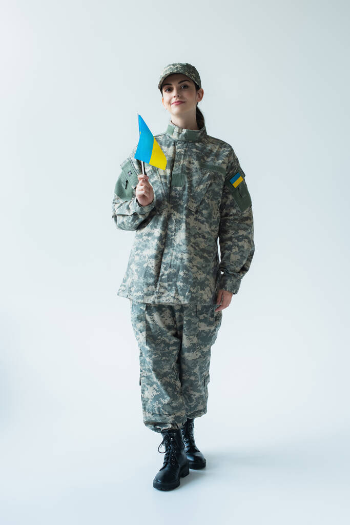 Smiling soldier in uniform holding ukrainian flag on grey background - Фото, зображення