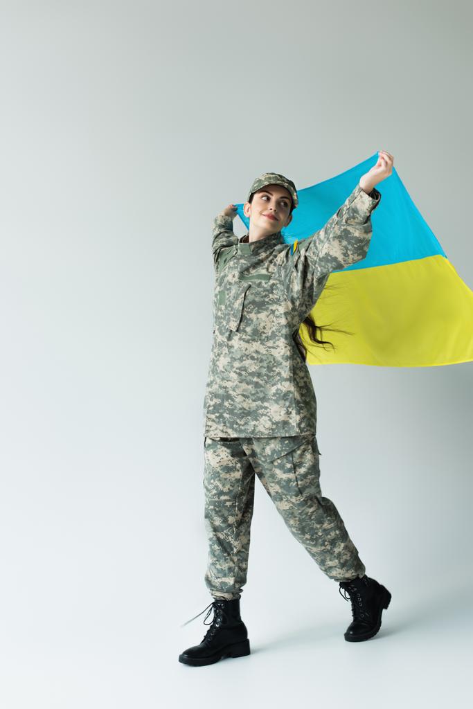 Full length of smiling soldier holding ukrainan flag while walking on grey background - Foto, Bild