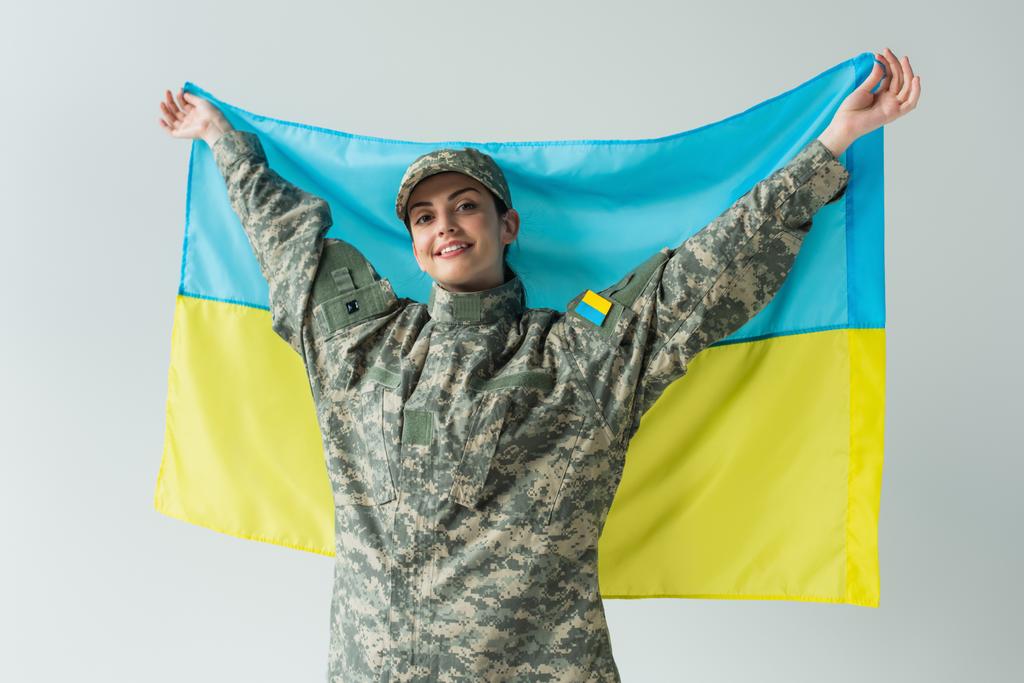 Smiling soldier in uniform holding ukrainian flag isolated on grey  - Photo, image