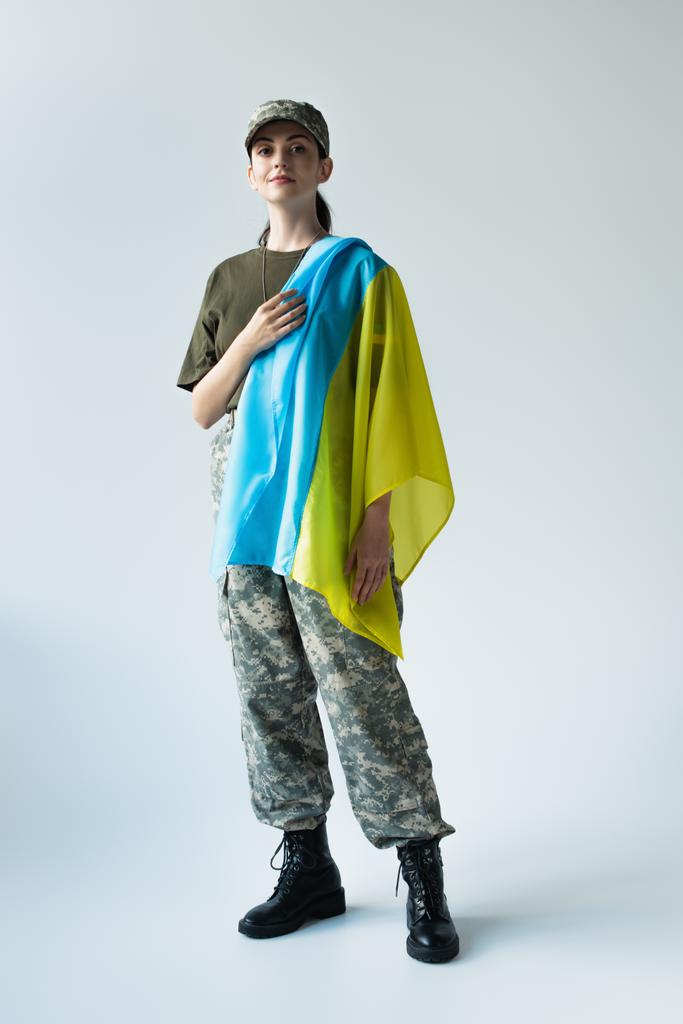 Full length of young soldier with ukrainian flag looking at camera on grey background - 写真・画像