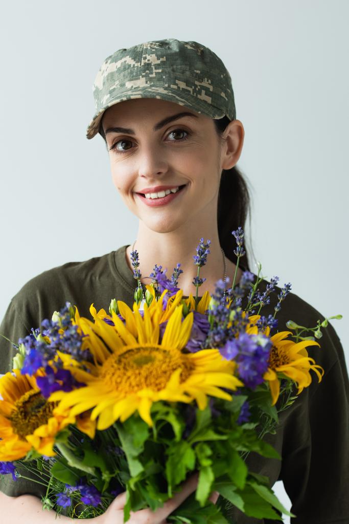 Portrait of positive servicewoman in cap holding blue and yellow flowers isolated on grey  - Fotografie, Obrázek