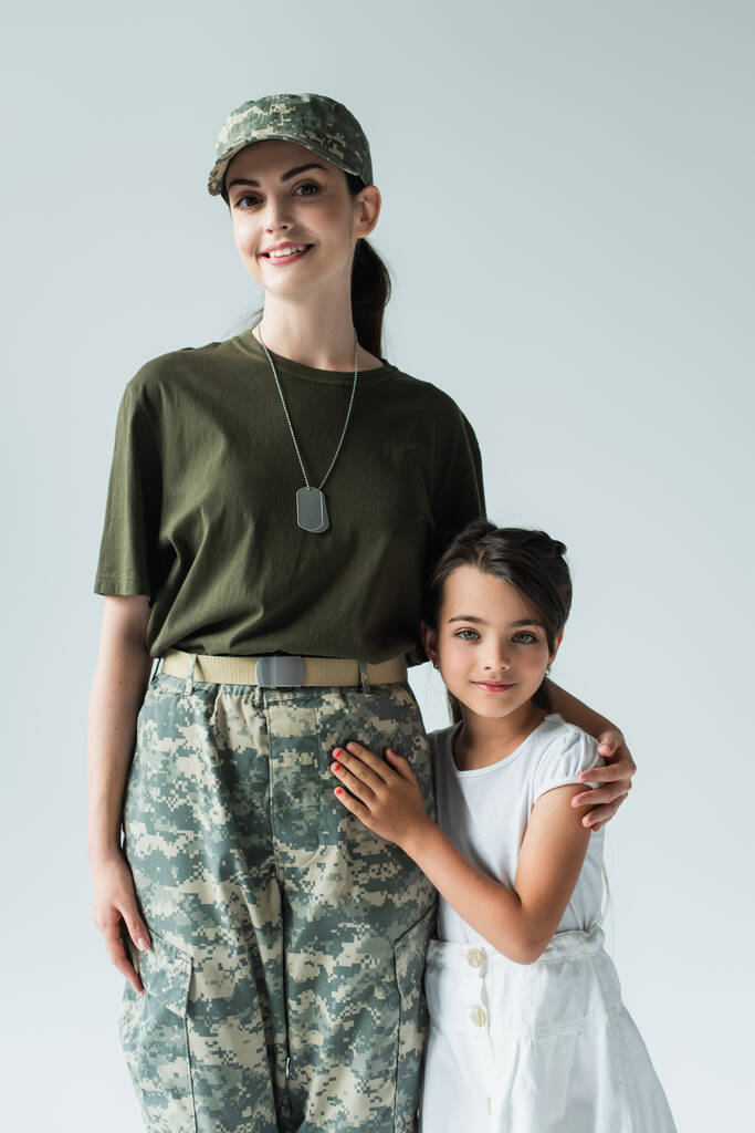 Smiling servicewoman in military uniform hugging child isolated on grey  - Foto, afbeelding