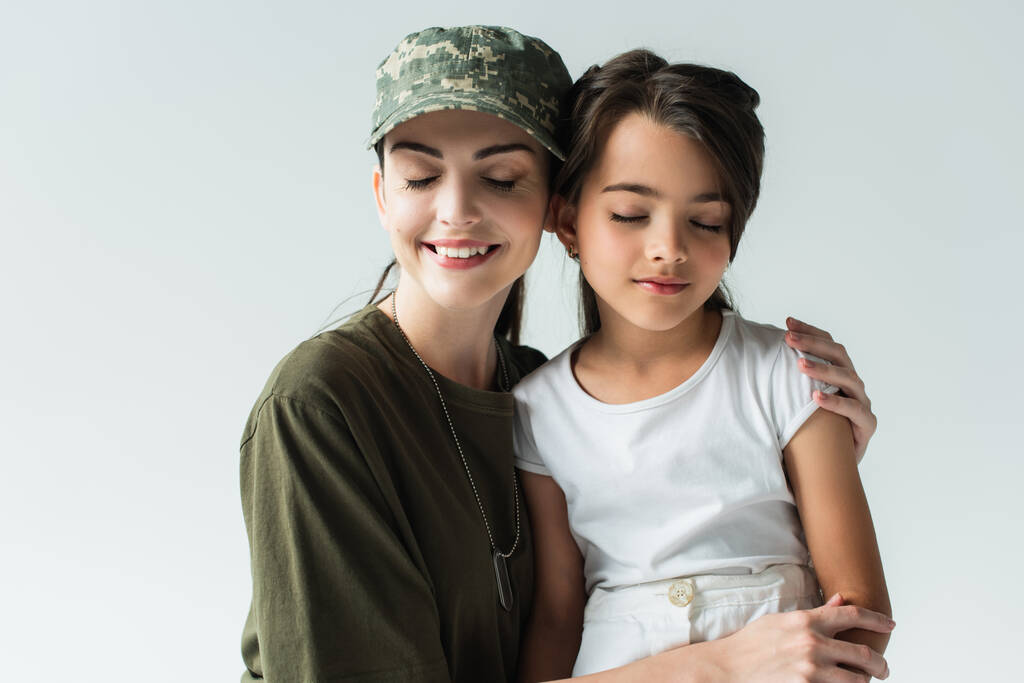 Mother in military uniform and daughter smiling and closing eyes isolated on grey  - Foto, Imagen