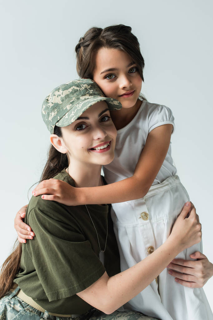 glimlachende moeder in militaire uniform kijken naar camera terwijl knuffelen kind geïsoleerd op grijs  - Foto, afbeelding