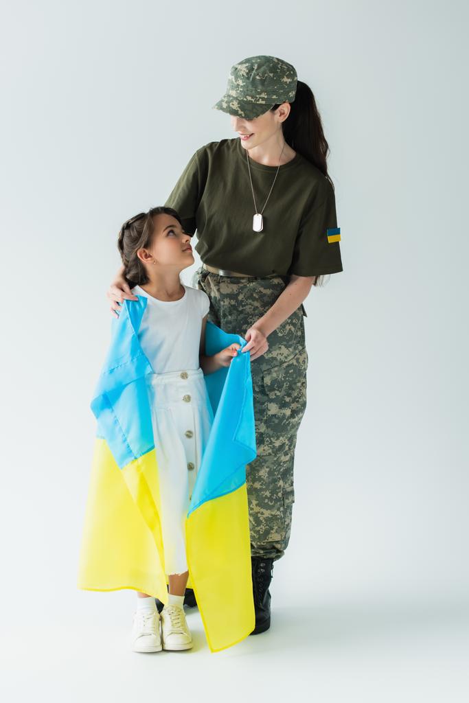 Kamuflaj üniformalı gülümseyen anne elinde Ukrayna bayrağı ve gri arka planda kızına sarılıyor. - Fotoğraf, Görsel