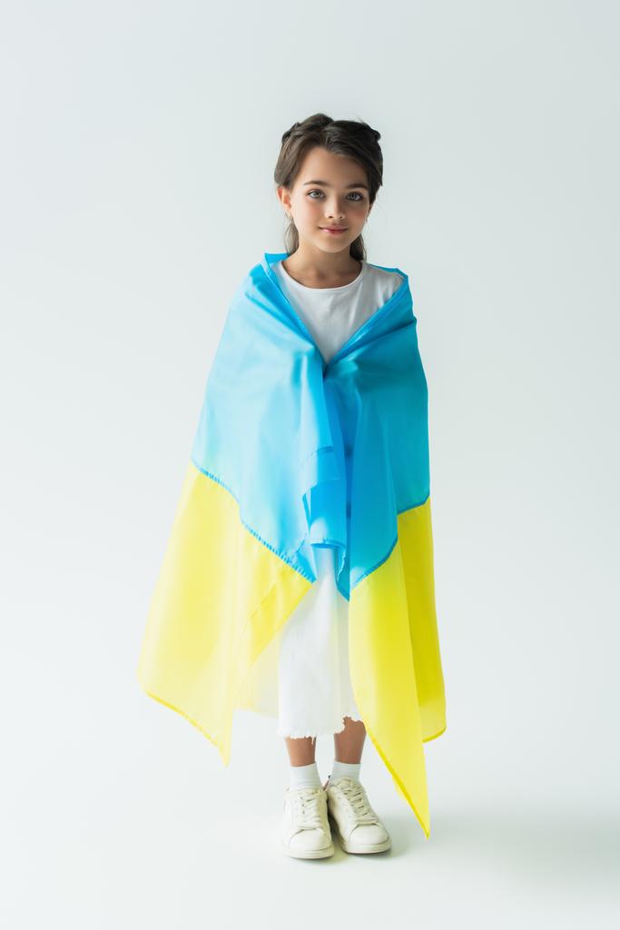 Smiling child covering with ukraining flag and standing on grey background - 写真・画像