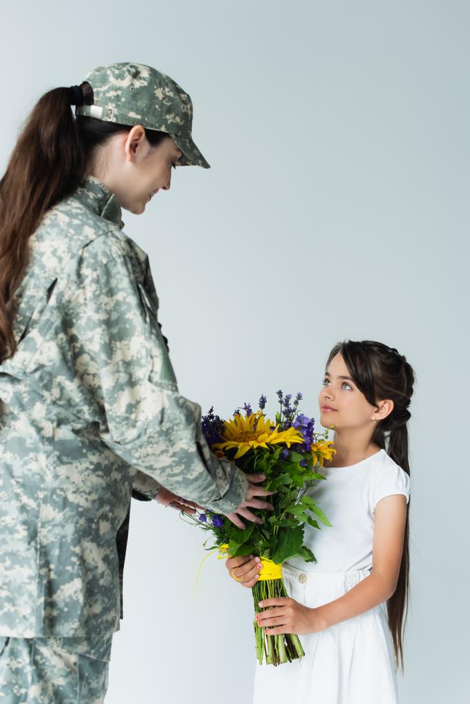 Kid giving blue and yellow bouquet to mother in camouflage uniform isolated on grey  - Foto, afbeelding