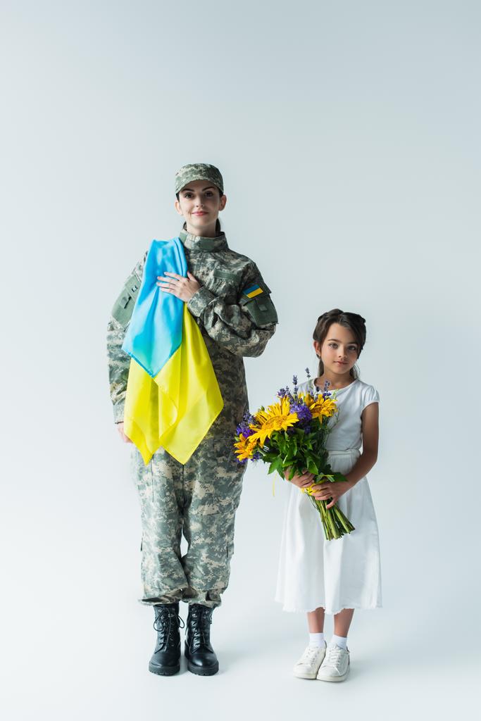 Żołnierz trzymający ukraińską flagę w pobliżu córki z bukietem na szarym tle - Zdjęcie, obraz