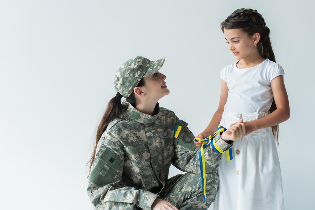 Kid držení modré a žluté stuhy a při pohledu na maminku ve vojenské uniformě izolované na šedé  - Fotografie, Obrázek