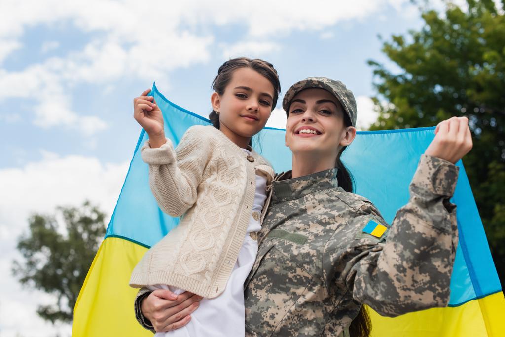 happy military woman with daughter and ukrainian flag looking at camera outdoors - Foto, imagen