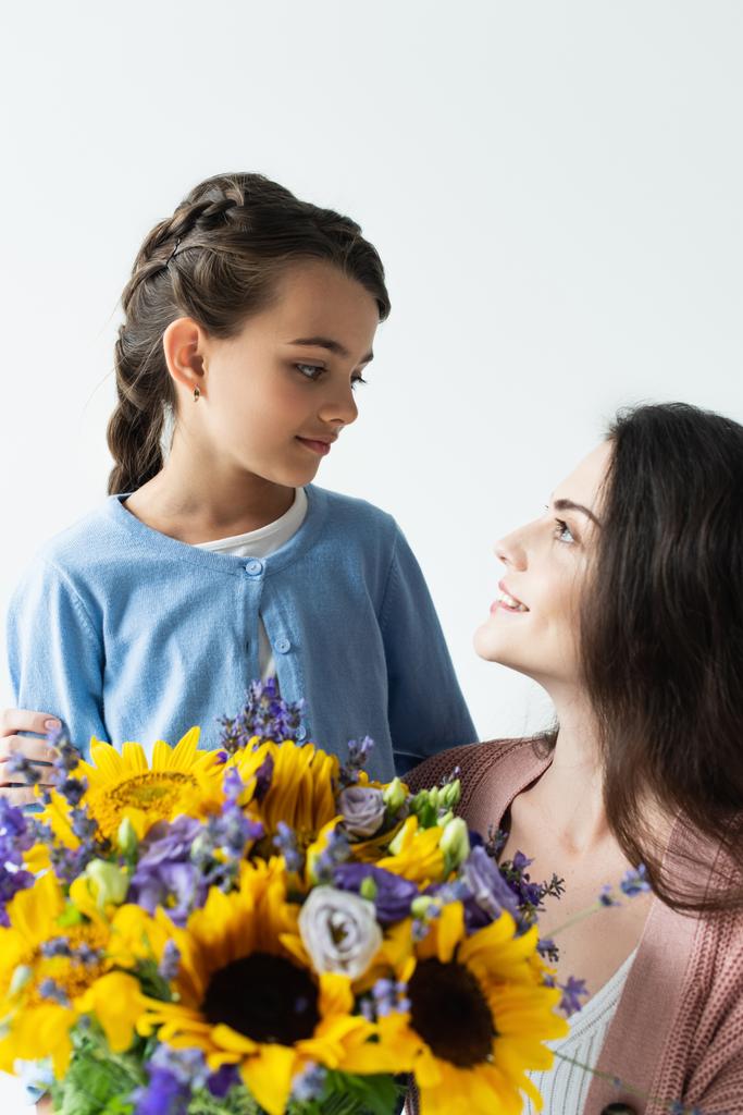 mom and daughter smiling at each other near blue and yellow flowers isolated on grey - 写真・画像