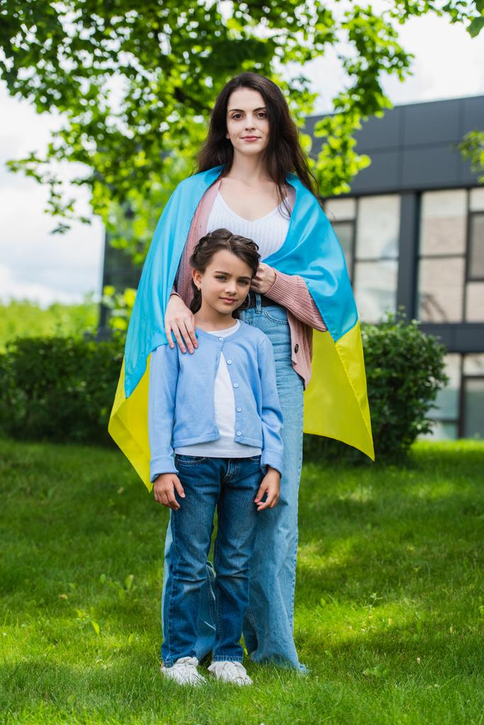 woman covered with ukrainian flag looking at camera near daughter in park - Zdjęcie, obraz