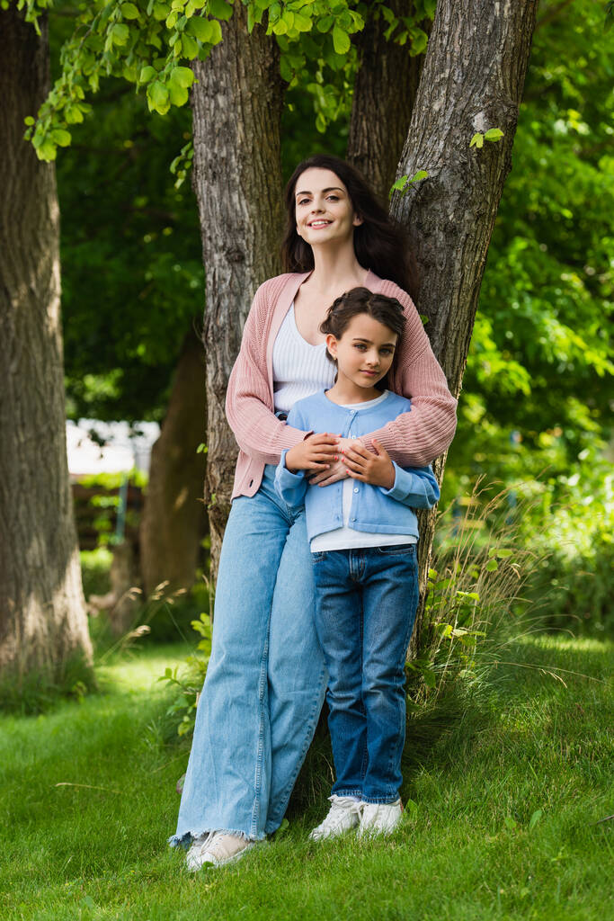full length of positive woman hugging daughter near trees in park - Foto, afbeelding