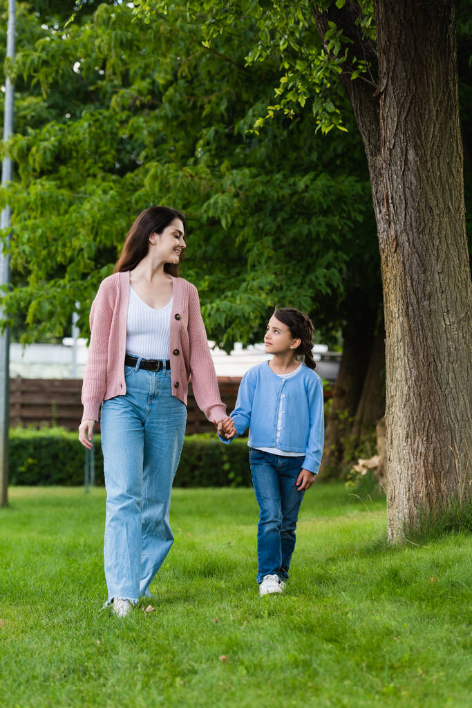 szczęśliwa kobieta i dziewczyna trzymając się za ręce i patrząc na siebie podczas spaceru w parku - Zdjęcie, obraz