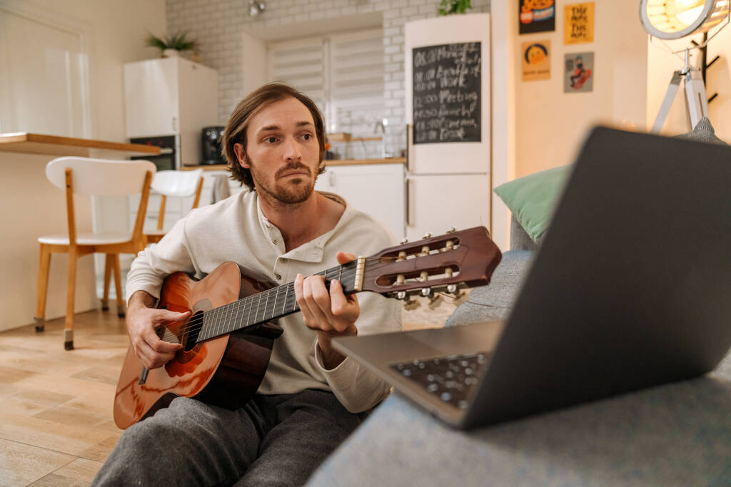 Імбирний чоловік грає на гітарі і використовує ноутбук, сидячи на підлозі вдома
 - Фото, зображення