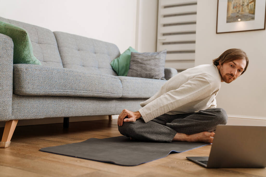 Gember man met behulp van laptop tijdens yoga praktijk op de vloer thuis - Foto, afbeelding