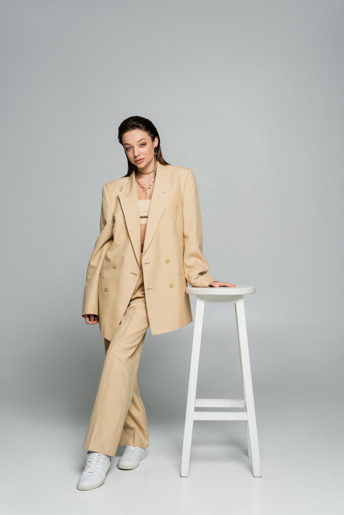 full length of pretty woman in beige blazer looking at camera while posing near high stool on grey - Foto, Imagen