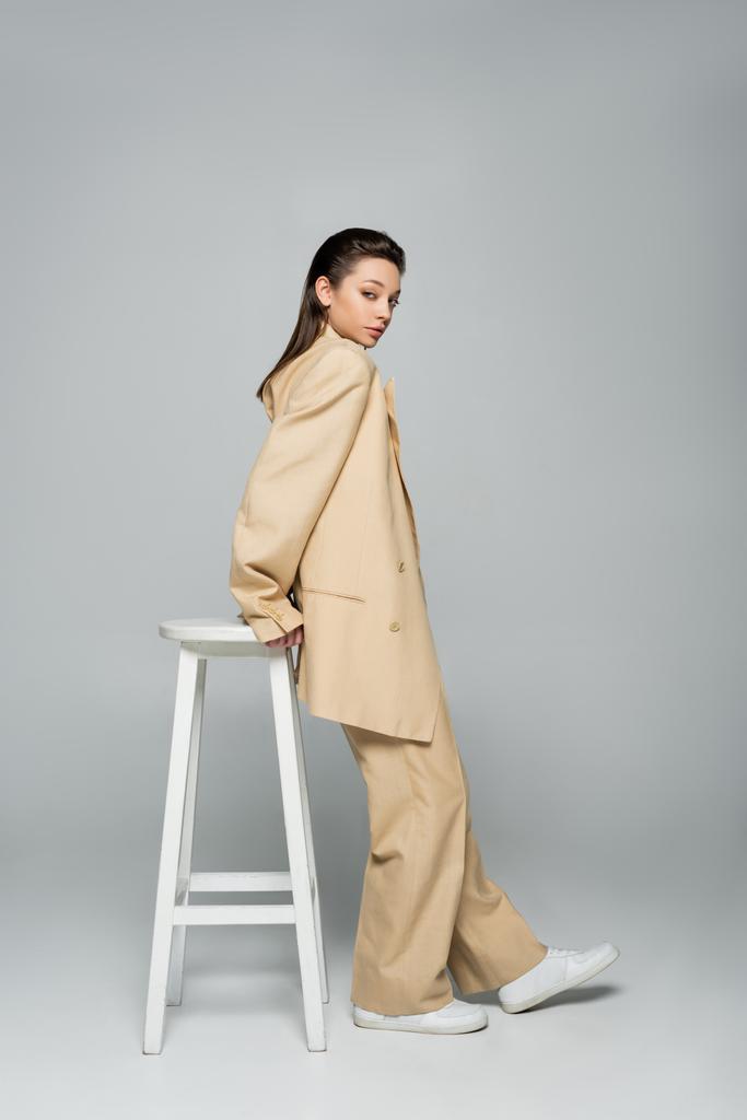 full length of young woman in beige suit looking at camera while posing near high stool on grey - Foto, afbeelding