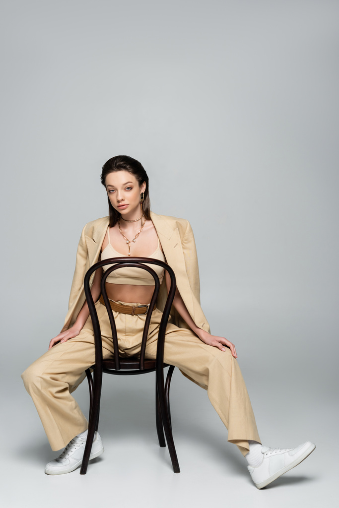 full length of young woman in beige outfit looking at camera while sitting on wooden chair on grey - Foto, Imagen