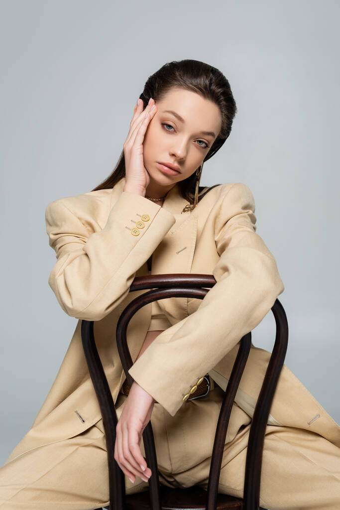 young woman in stylish outfit looking at camera while sitting on chair isolated on grey - Fotografie, Obrázek