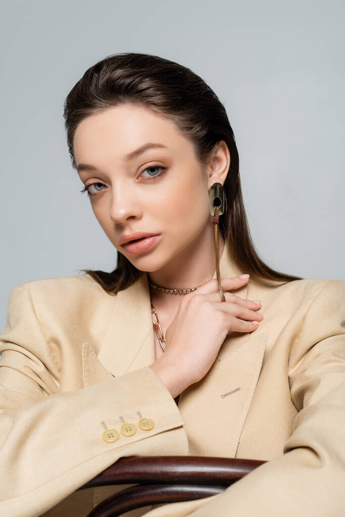 young woman in stylish outfit and golden earring looking at camera while posing isolated on grey - Foto, afbeelding