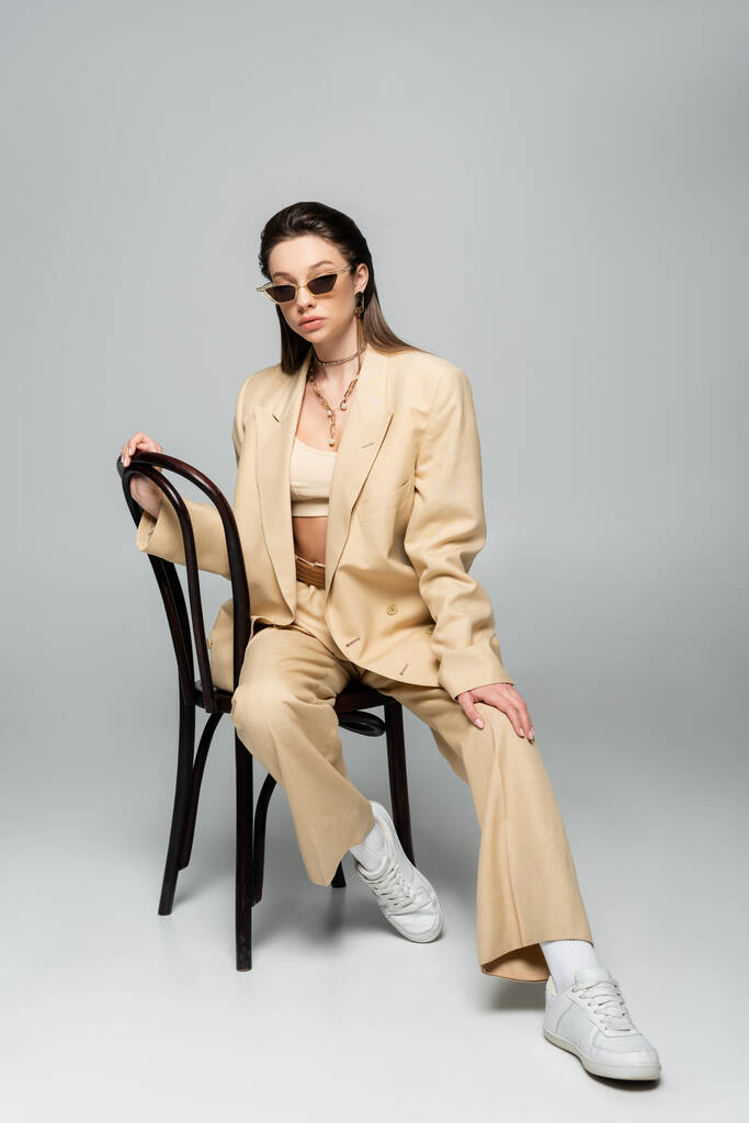full length of young woman in stylish beige outfit and trendy sunglasses sitting on wooden chair on grey - Zdjęcie, obraz