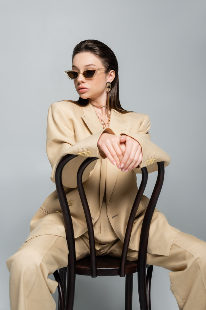 young woman in stylish beige outfit and sunglasses sitting on wooden chair isolated on grey - Fotoğraf, Görsel