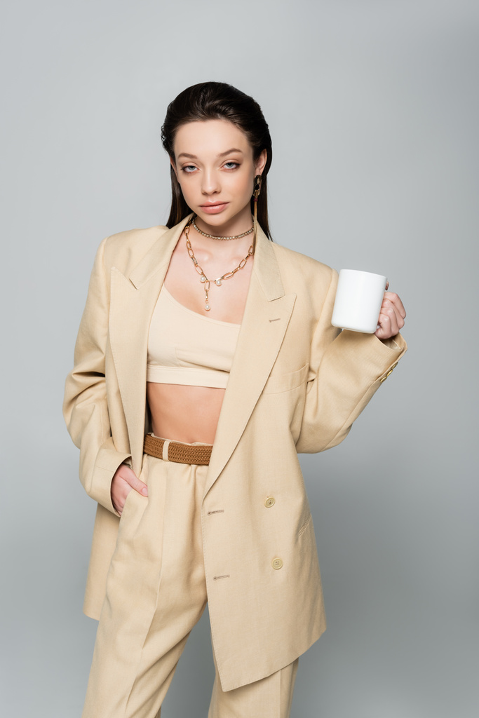 brunette woman in stylish beige suit holding cup of coffee and posing with hand in pocket isolated on grey - Fotoğraf, Görsel