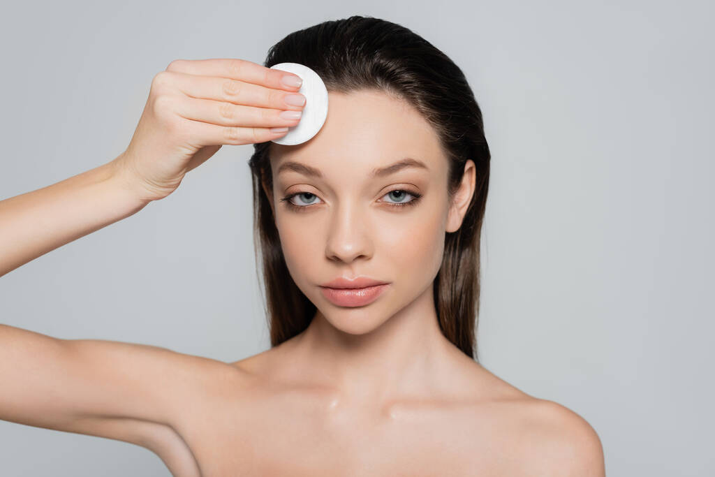 young woman removing makeup on forehead with soft cotton pad isolated on grey - Foto, imagen