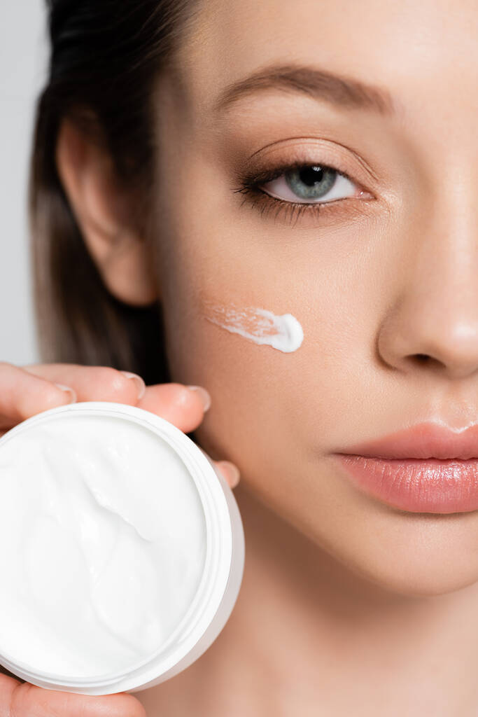 close up view of cropped young woman with cosmetic cream on cheek holding container isolated on grey - Foto, Imagem
