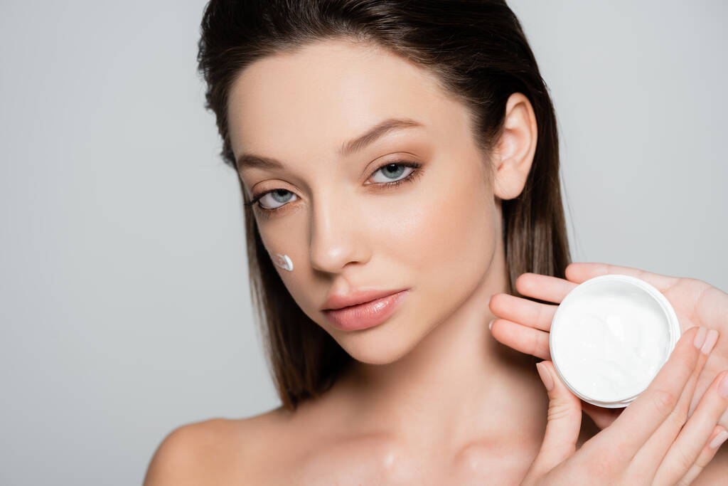 brunette woman with bare shoulders and cosmetic cream on cheeks holding container isolated on grey - Foto, Imagem