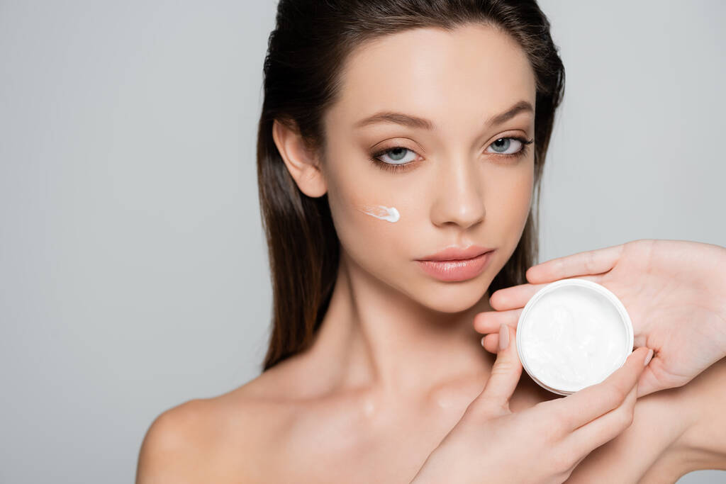 young woman with cosmetic cream on cheeks holding container isolated on grey - Foto, Imagem