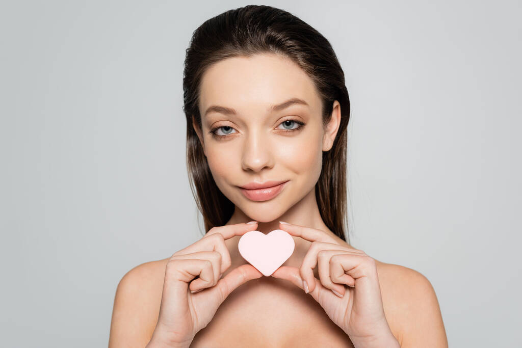 smiling young woman holding heart-shaped sponge near face isolated on grey - Fotó, kép