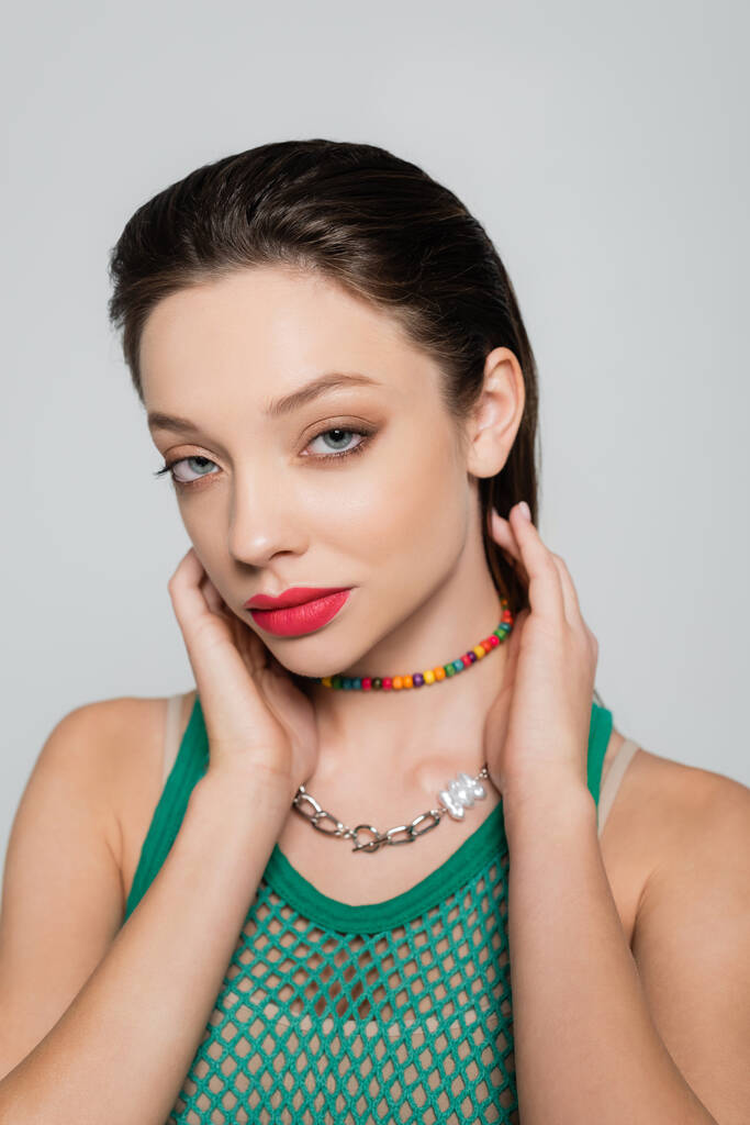 stylish model in green tank top and colorful necklace looking at camera isolated on grey - Valokuva, kuva
