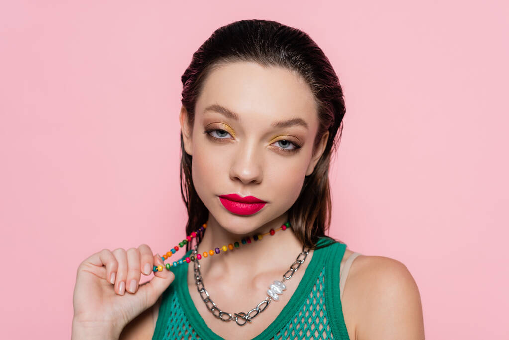 young brunette woman with bright makeup pulling beads necklace while looking at camera isolated on pink  - Foto, Bild
