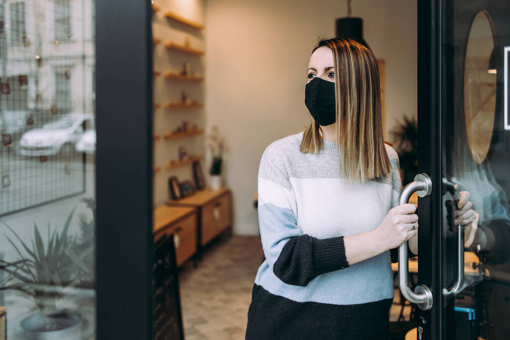 オープンワークショップのドアに立って顔の保護マスクを持つ若く魅力的な女性アーティストと外を見る。彼女は自信があり、真剣だ。ビジネスコンセプトのためにオープンCovid-19. - 写真・画像
