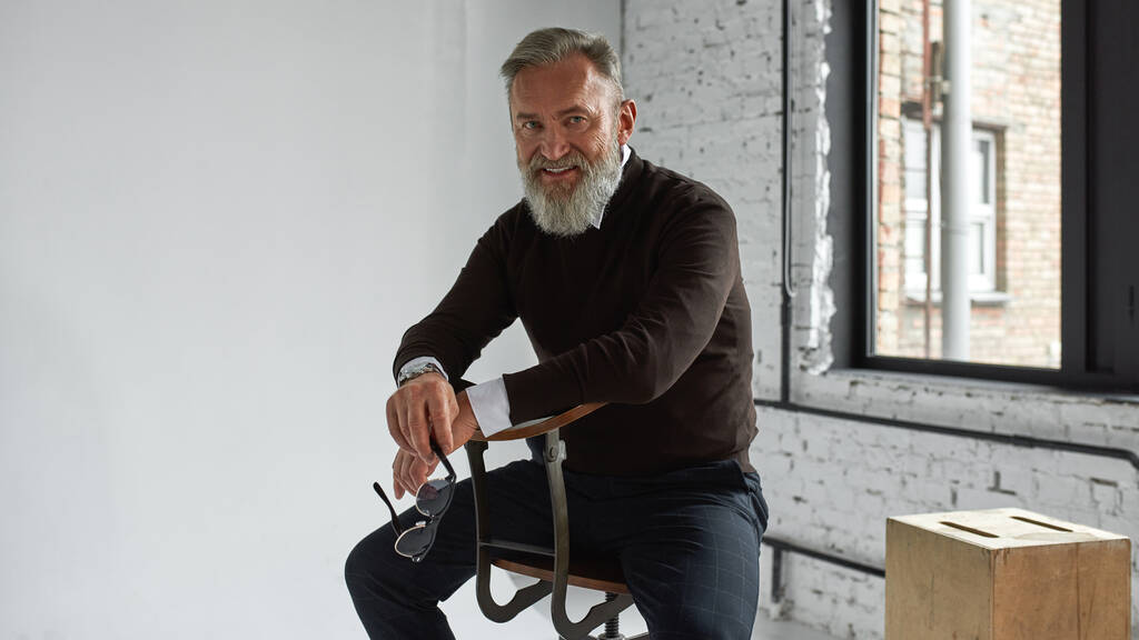 Grey hair confident caucasian man sitting on chair and looking at camera in spacious flat. Fashionable bearded male with glasses wearing casual clothes. Concept of modern elderly male lifestyle - Foto, Imagem