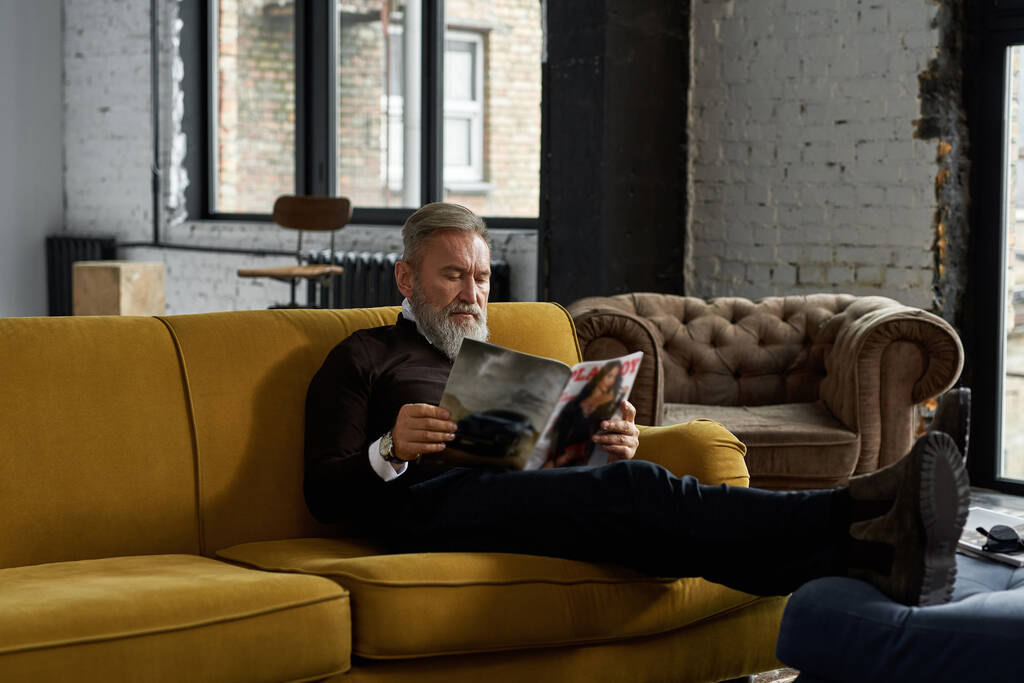Elderly focused caucasian man reading journal on sofa at home. Trendy bearded pensioner wearing casual clothes. Modern senior male lifestyle. Domestic rest and leisure. Spacious apertment interior - Foto, immagini